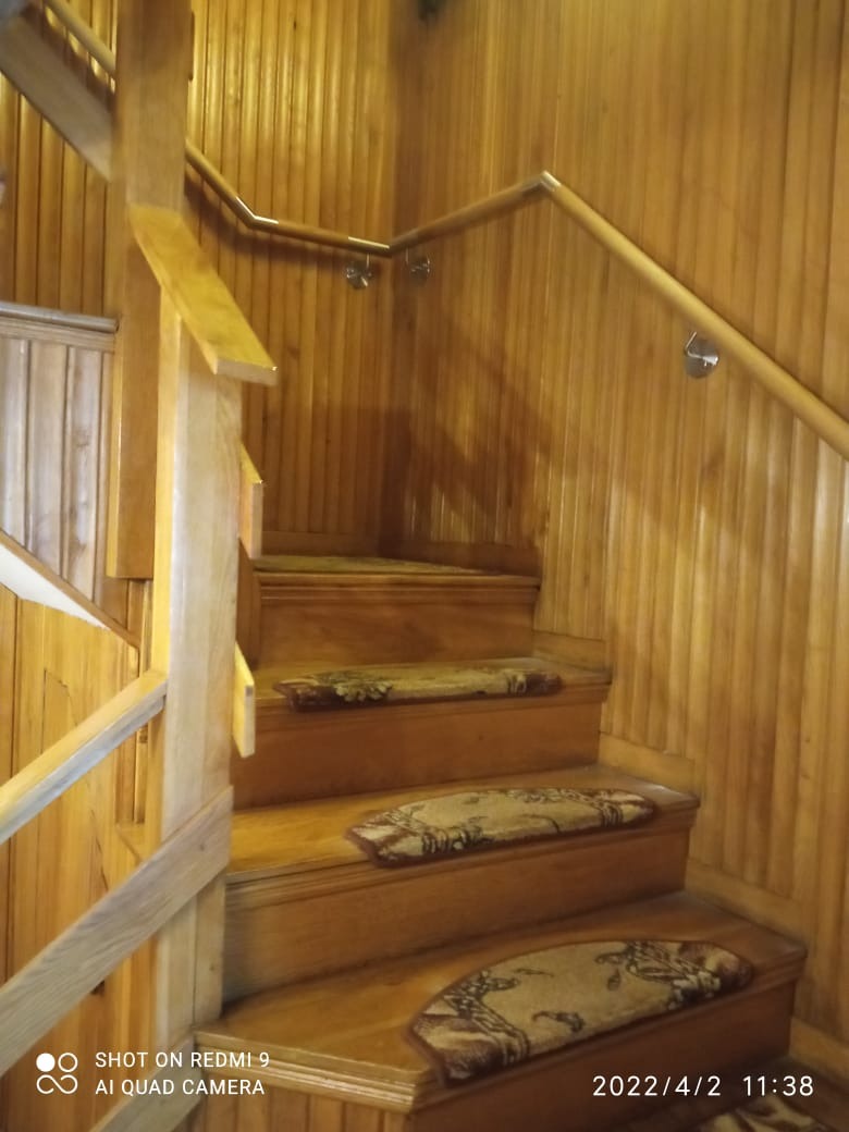Drewniane schody z metalową poręczą na tle boazerii, widoczne małe dywaniki na stopniach.