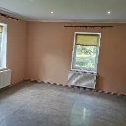 Pomalowane na brzoskwiniowo wnętrze pokoju z dwoma oknami z roletami, grzejnikami pod oknami i szarą podłogą z płytek imitujących kamień.