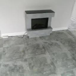 Nowoczesny, elektryczny kominek obudowany szarą płytą imitującą beton, na tle szarej podłogi z płytek i białych ścian.