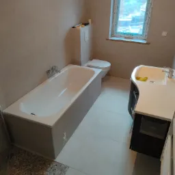 Wyremontowana łazienka w beżowych płytkach z wanną, umywalką z szafką, toaletą i oknem zabezpieczonym folią.