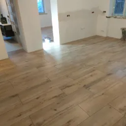 Ułożona podłoga z płytek imitujących drewno w jasnym odcieniu, widok na białe ściany z przygotowanymi punktami elektrycznymi i otwarte przejście do innego pomieszczenia z oknem i grzejnikiem.