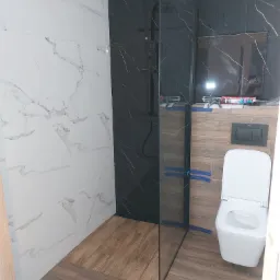 Nowoczesna łazienka z czarną kabiną prysznicową typu walk-in, białymi marmurkowymi płytkami ściennymi oraz podłogą imitującą drewno. Widoczna toaleta i akcesoria montażowe na półce.