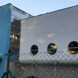 Nowoczesny budynek z niebieską i białą elewacją, okrągłymi oknami i zielonymi rurami wentylacyjnymi na zewnątrz, widok zza ogrodzenia z siatki.