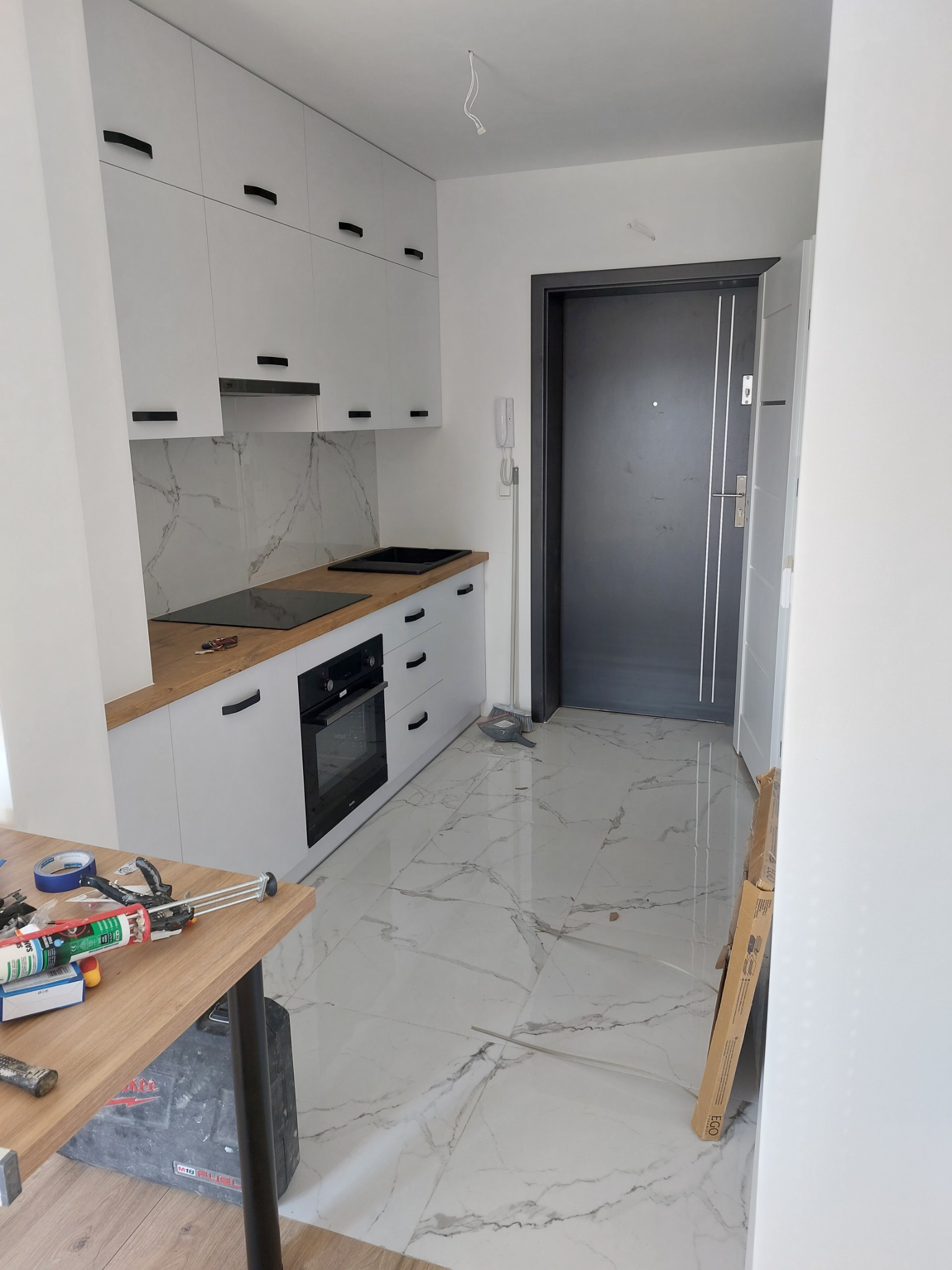 Nowoczesna kuchnia z białymi szafkami, drewnianym blatem, marmurową ścianą i podłogą, widok na czarne drzwi wejściowe, narzędzia na stole sugerują trwające prace wykończeniowe.