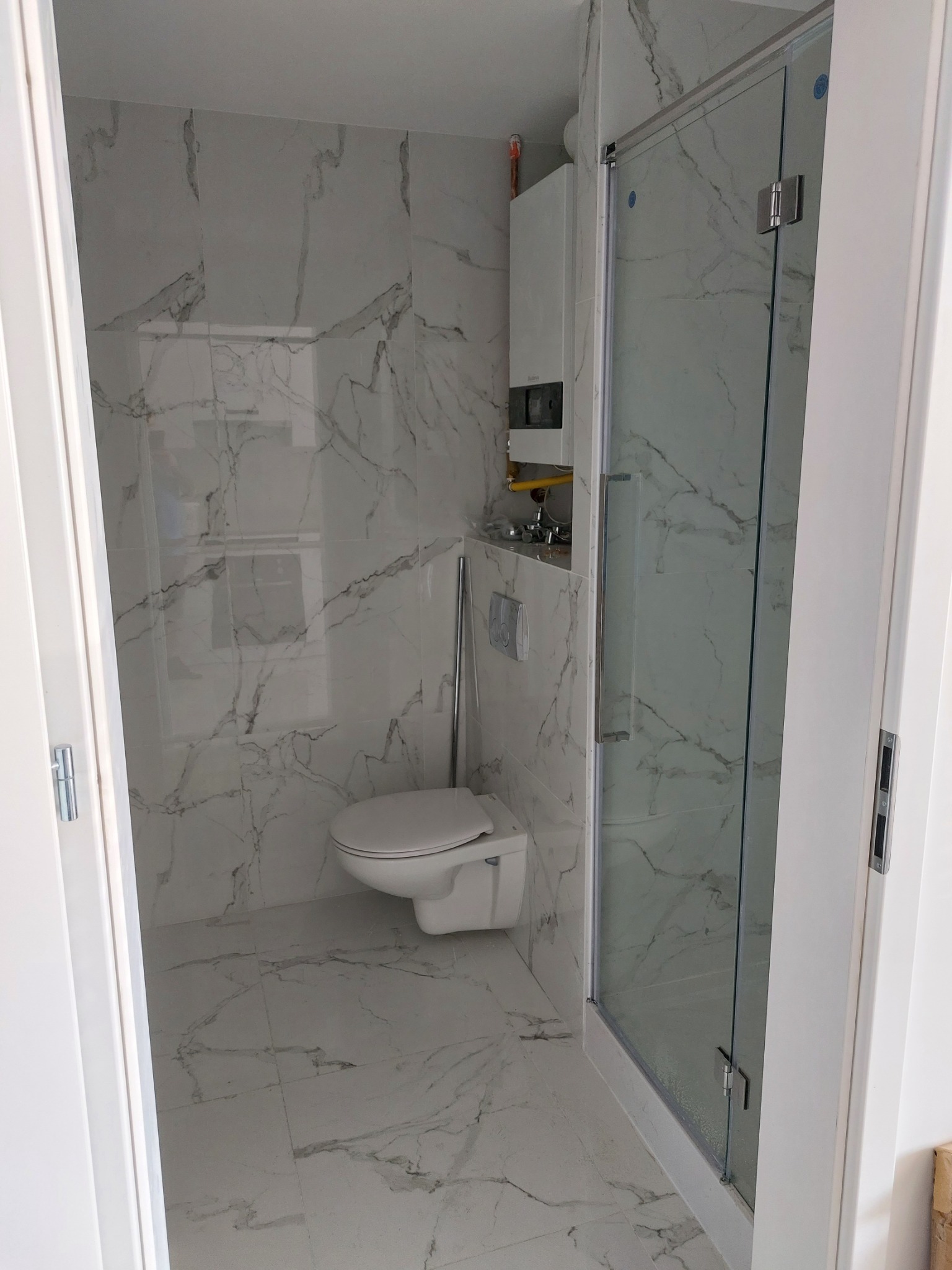 Nowoczesna łazienka wykończona białymi płytkami imitującymi marmur, z kabiną prysznicową ze szklanymi drzwiami i podwieszaną toaletą.