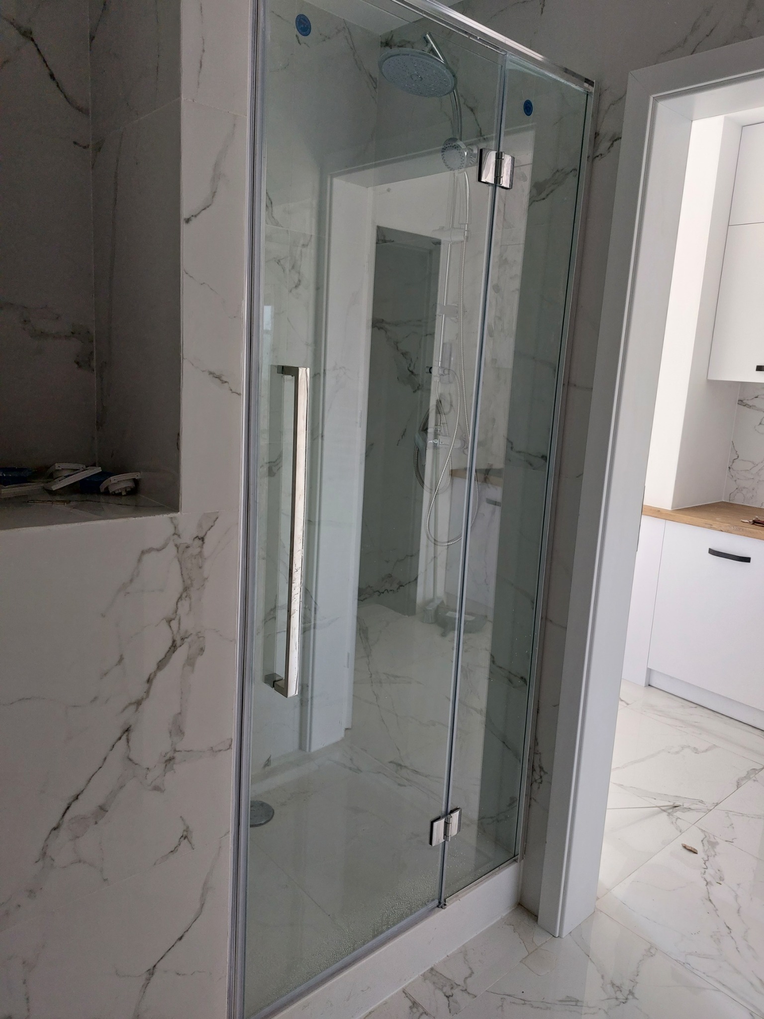 Szklana kabina prysznicowa z chromowanymi okuciami i uchwytem, zamontowana w łazience wykończonej płytkami imitującymi marmur, widoczny odpływ liniowy w brodziku.