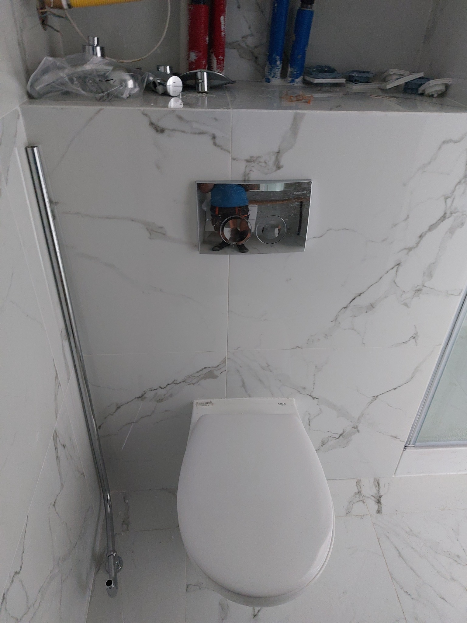 Biała toaleta wisząca z chromowanym przyciskiem spłukującym na tle marmurowych płytek, widoczne elementy instalacji wodnej i odbicie osoby w przycisku.