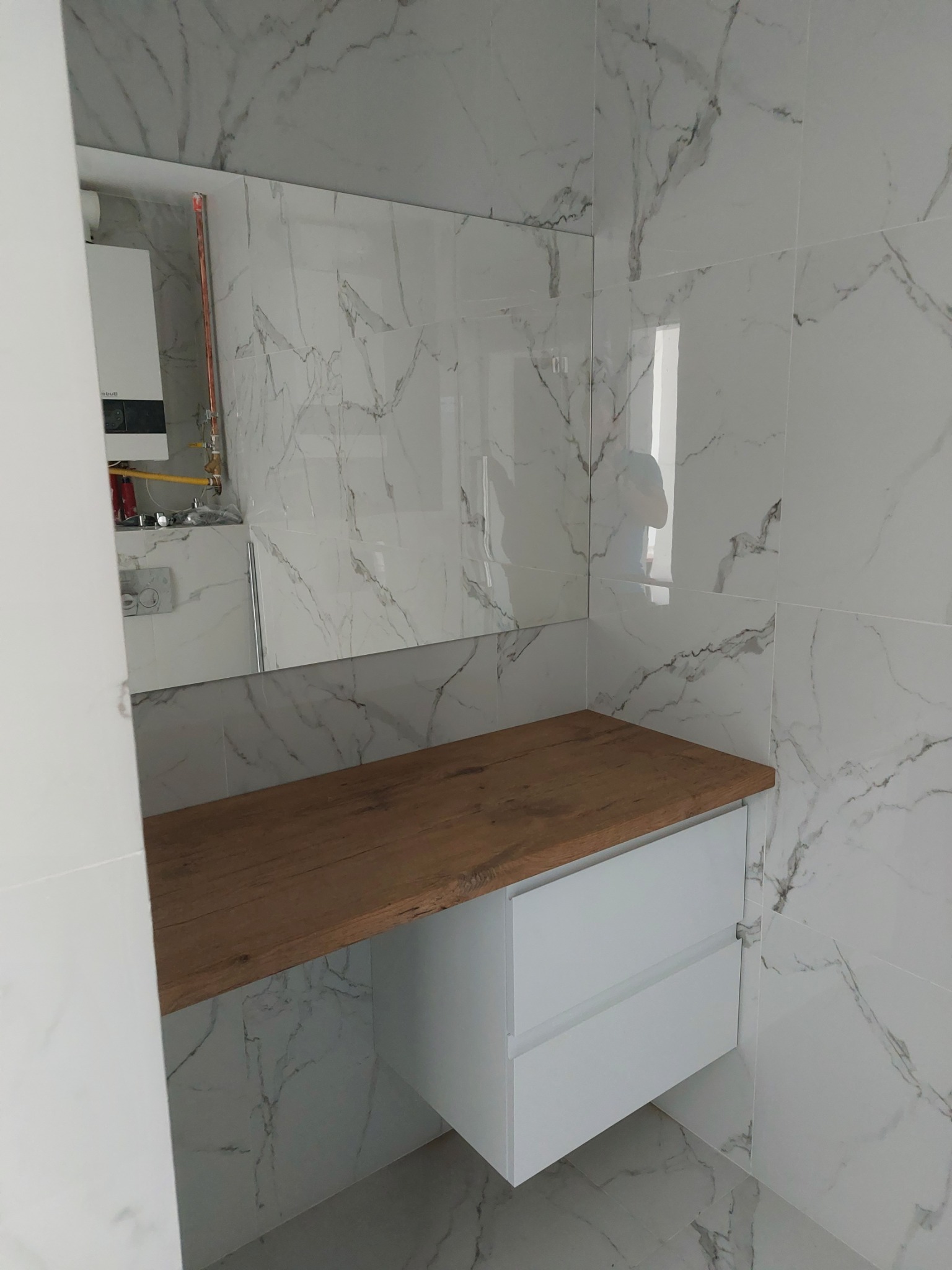 Nowoczesna łazienka z białymi płytkami imitującymi marmur, drewnianym blatem i białą szafką z dwiema szufladami. W lustrze odbija się bojler i instalacja.