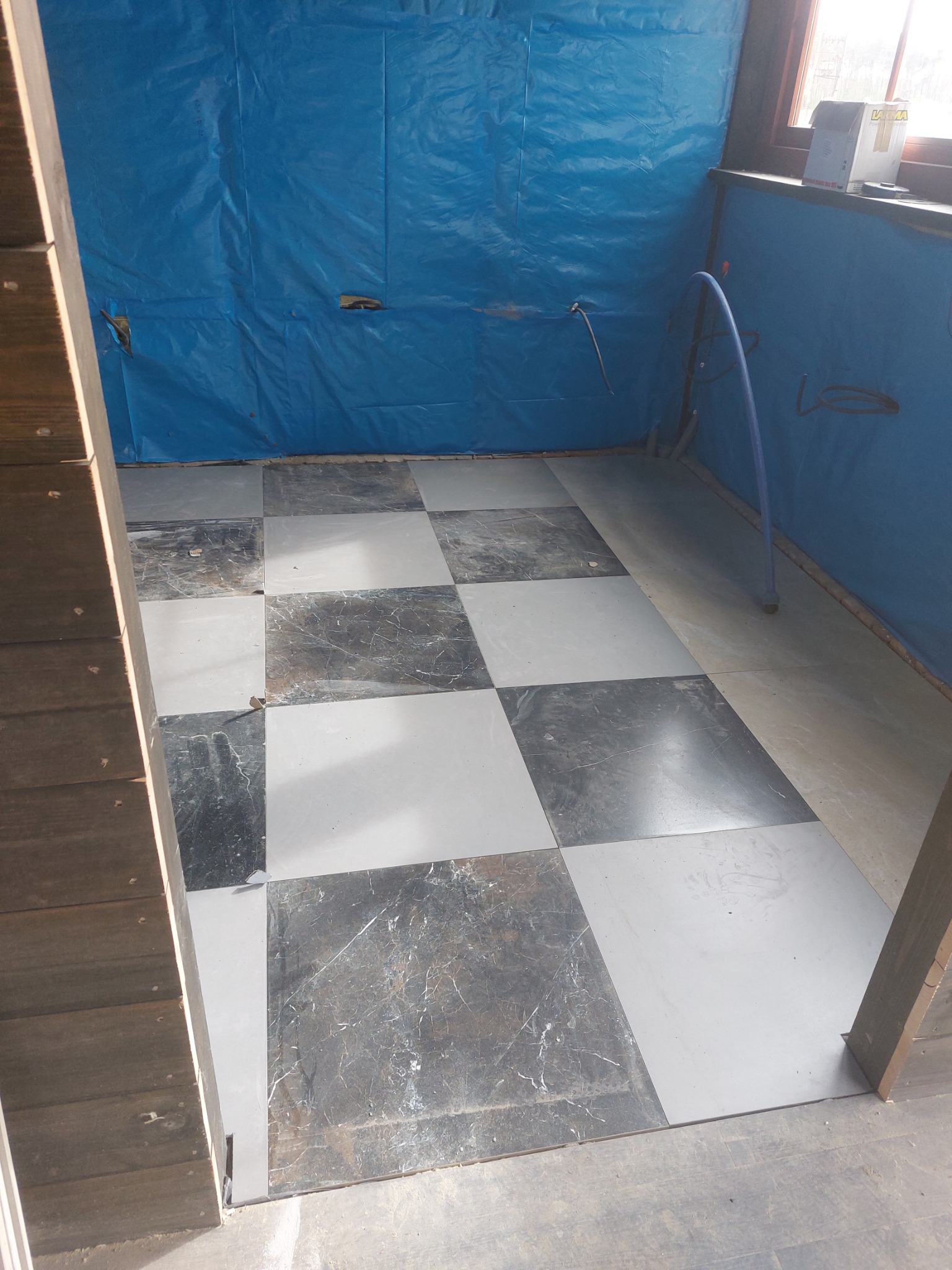Ułożona podłoga z szaro-białych płytek w szachownicę, ściany zabezpieczone niebieską folią malarską, widoczne rury instalacji.