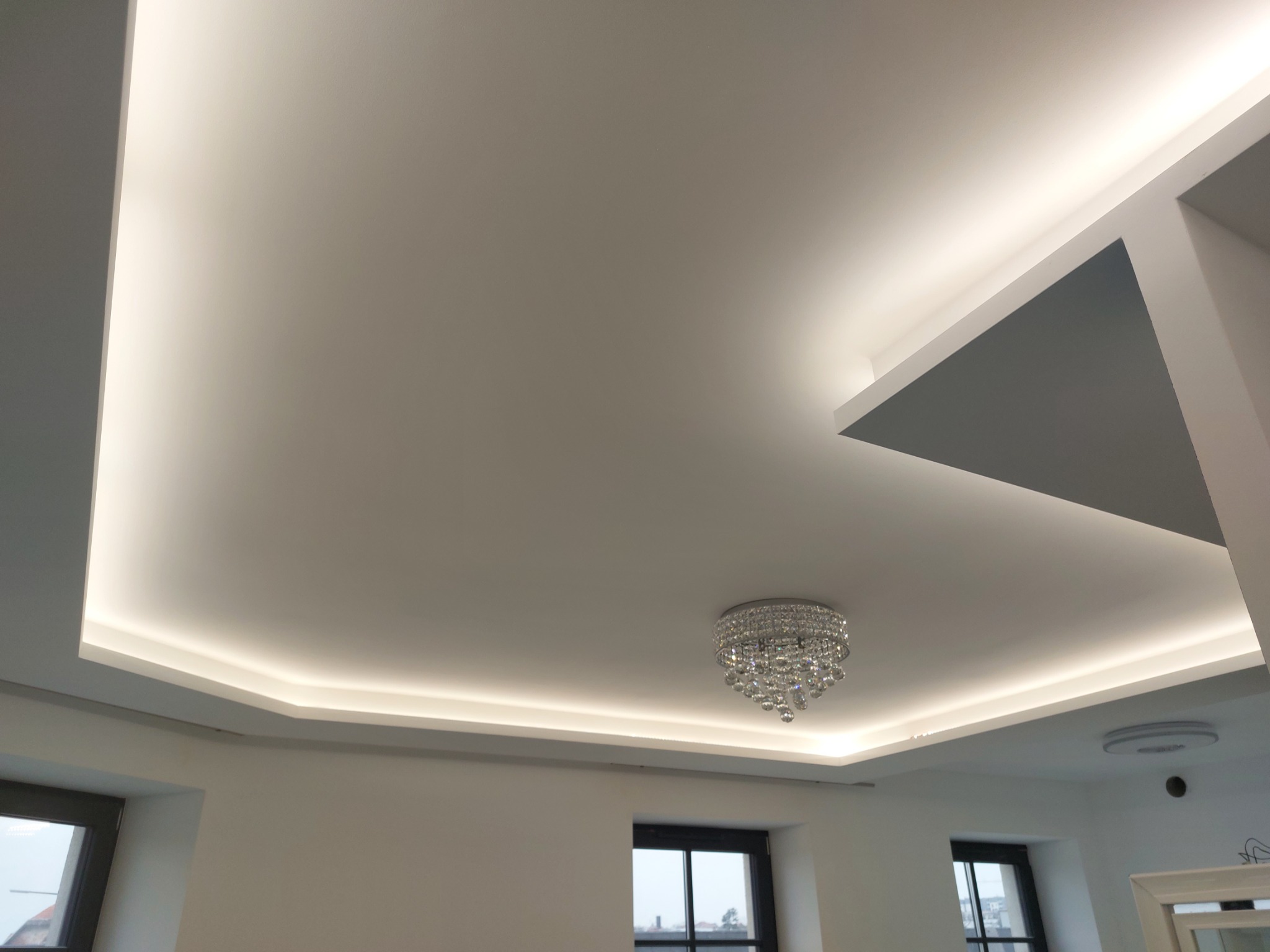 Biały sufit podwieszany z oświetleniem LED i kryształowym żyrandolem w nowoczesnym wnętrzu, widoczne okna z ciemnymi ramami.