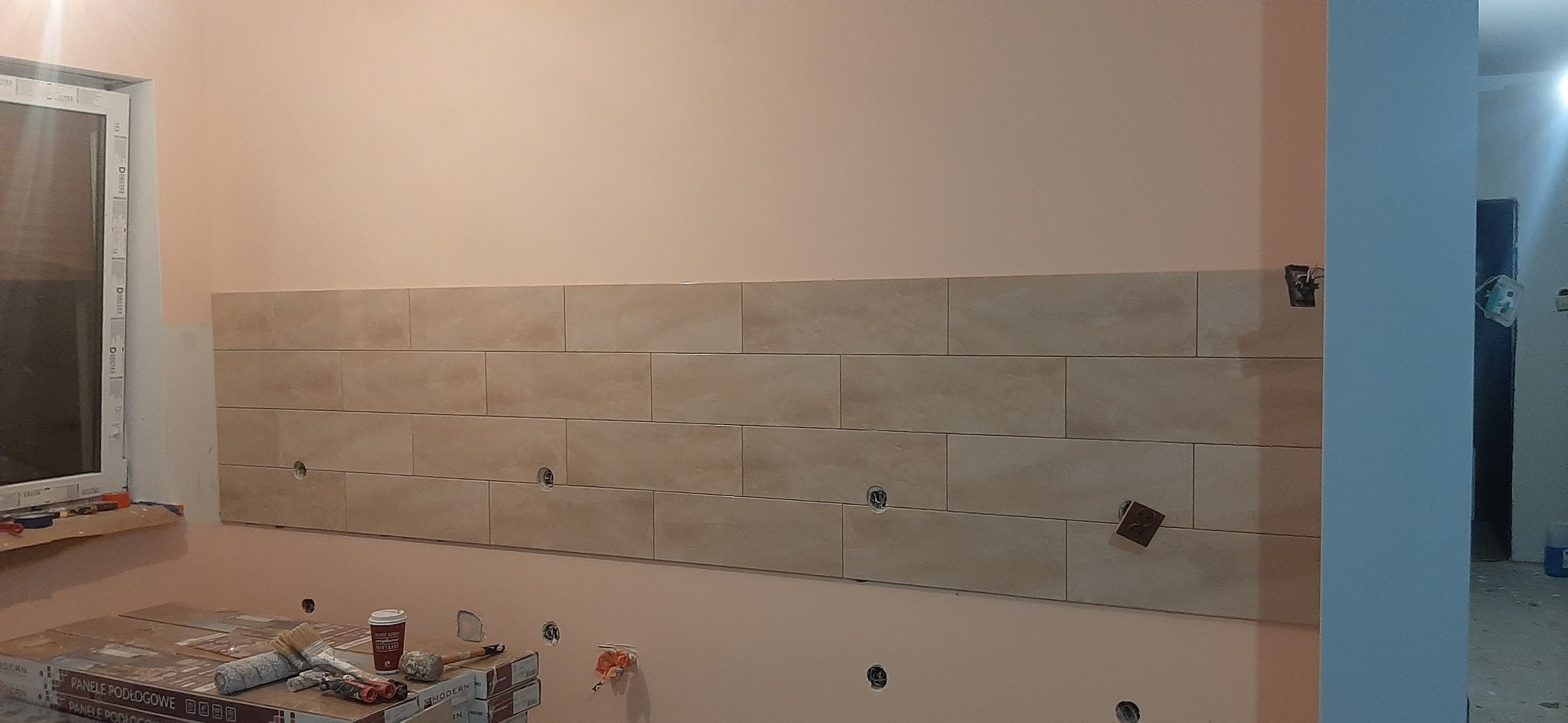 Ściana z beżowymi, prostokątnymi płytkami ceramicznymi ułożonymi poziomo, z widocznymi otworami na gniazdka elektryczne i elementami narzędzi glazurniczych na podłodze.