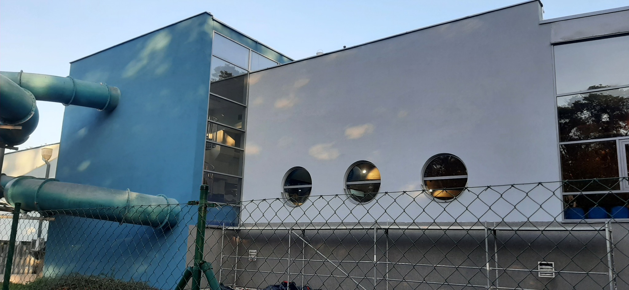 Nowoczesny budynek z niebieską i białą elewacją, okrągłymi oknami i zielonymi rurami wentylacyjnymi na zewnątrz, widok zza ogrodzenia z siatki.