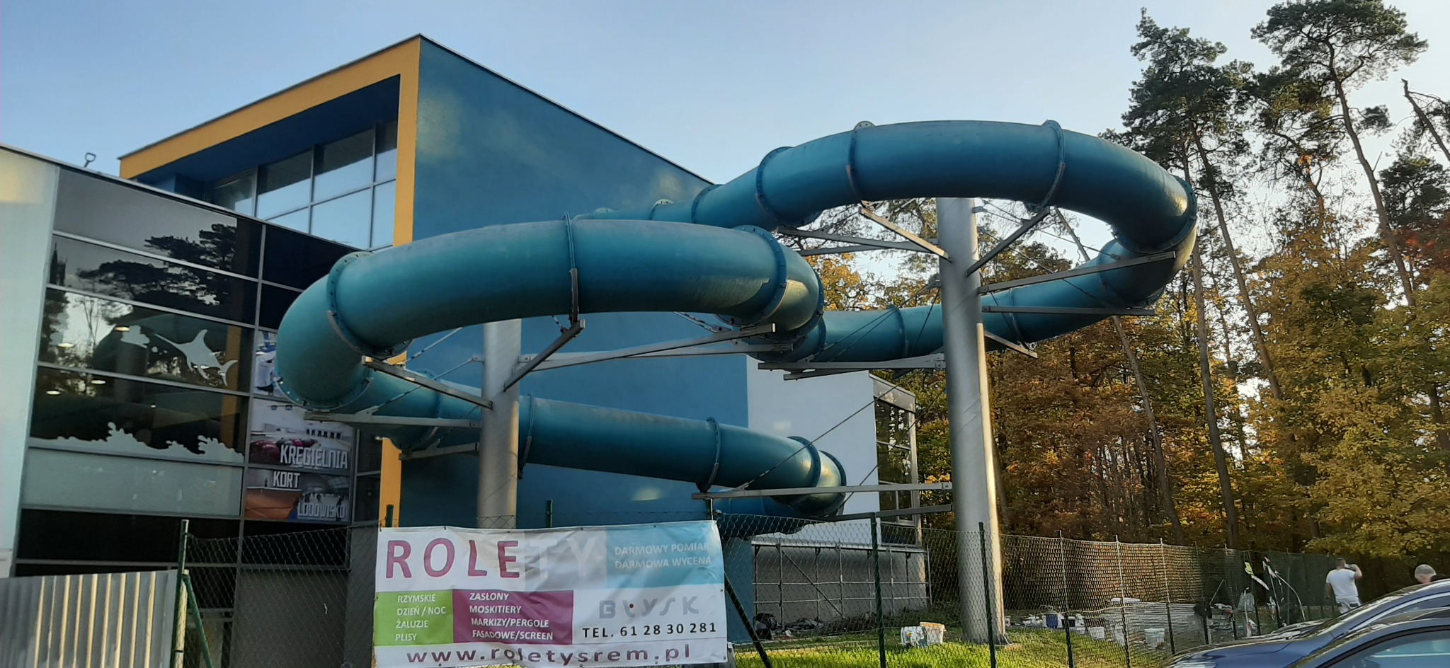 Błękitny zjeżdżalnia wodna z rur na tle budynku z żółtymi akcentami architektonicznymi i jesiennych drzew, widoczny baner reklamowy firmy Rolety Srem.