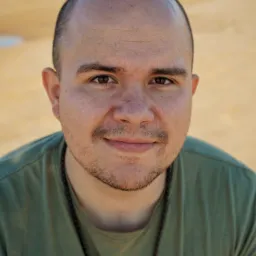 Portret mężczyzny w zielonej koszulce z delikatnym uśmiechem, na rozmytym, jasnym tle.