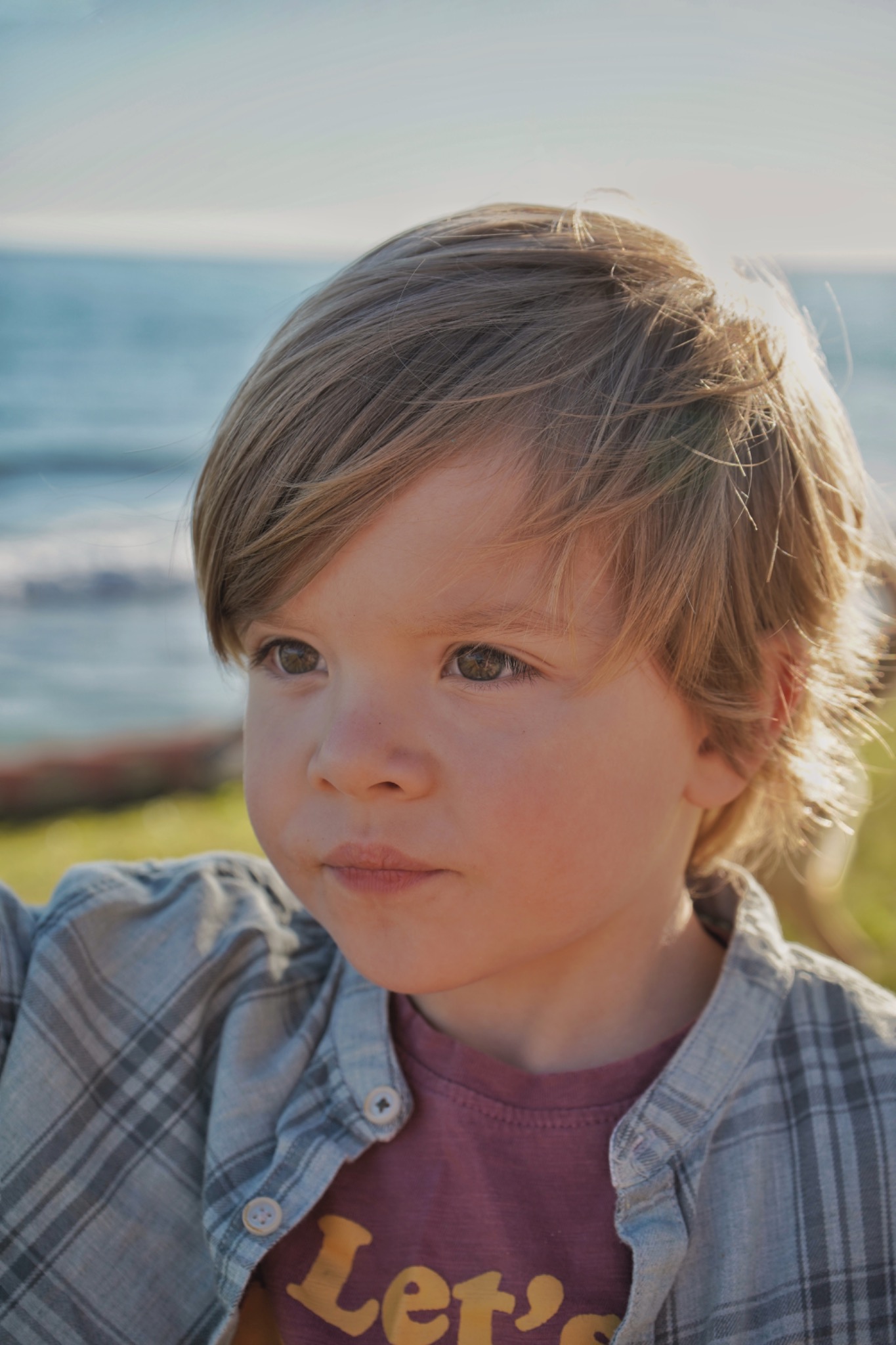 Portret małego chłopca z blond włosami i brązowymi oczami, ubrany w koszulę w kratę, z widokiem na morze w tle.
