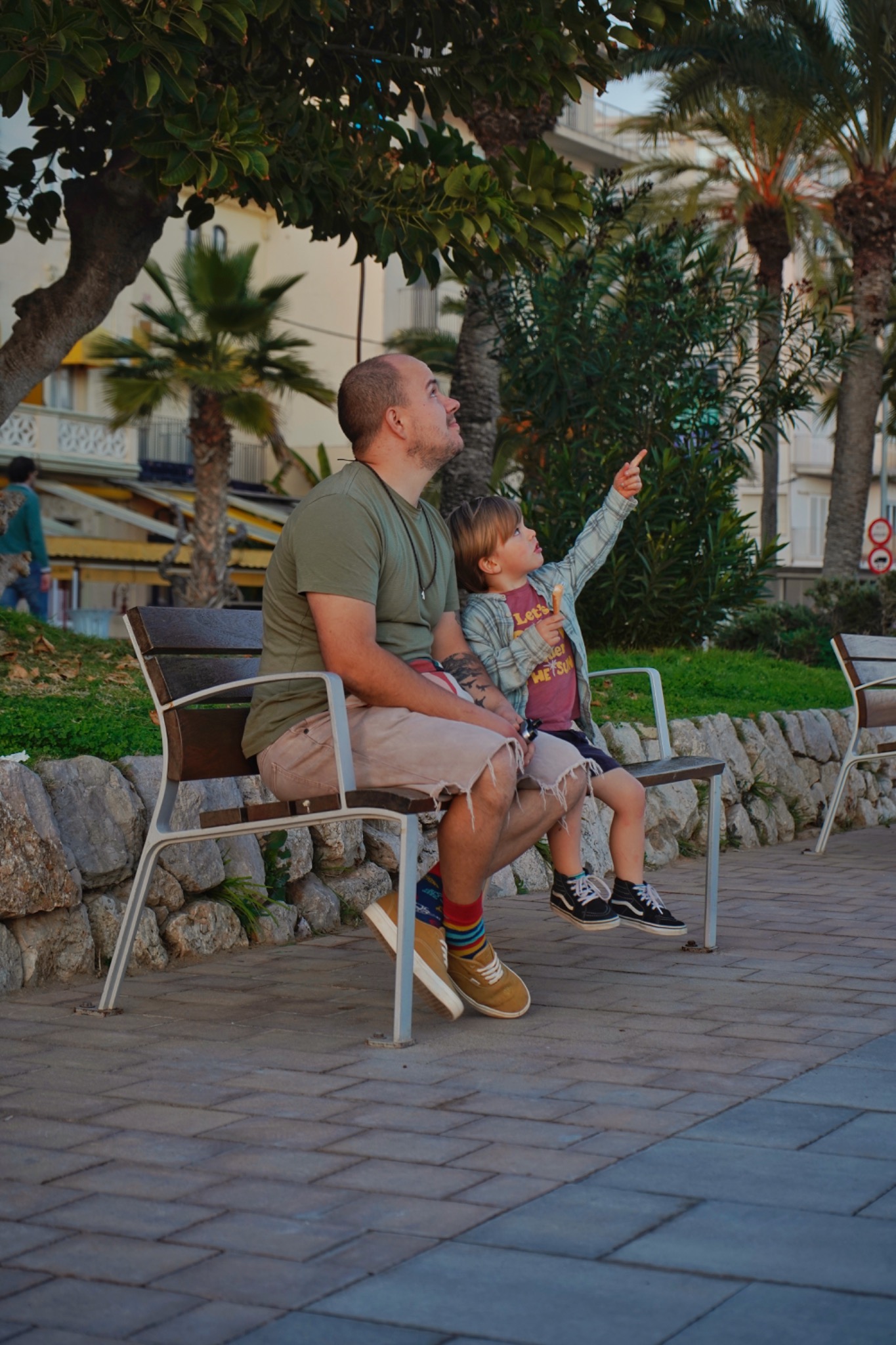 Ojciec z synem siedzą na ławce w parku, obaj patrzą w górę; chłopiec wskazuje palcem, trzymając w drugiej ręce loda.