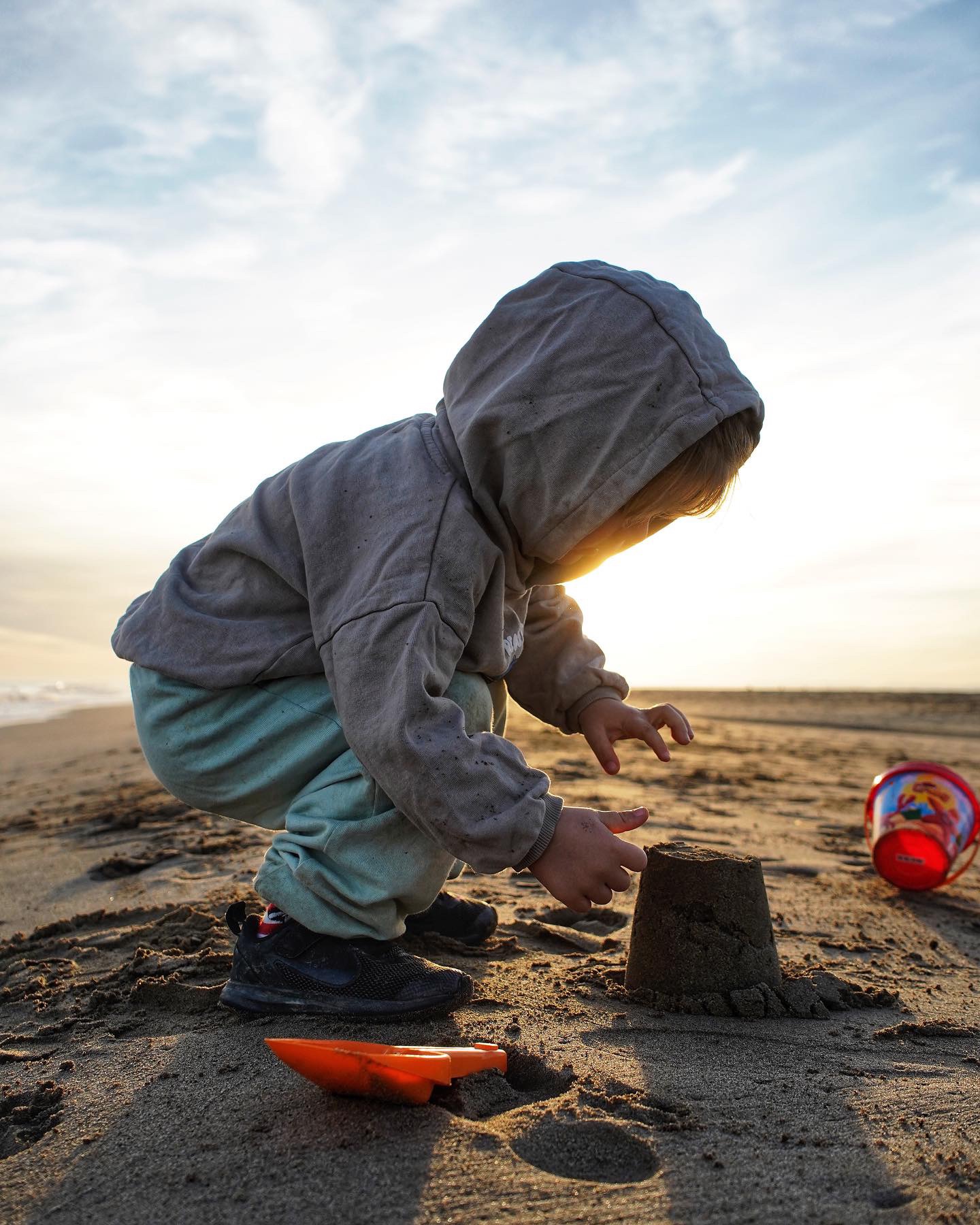 Dziecko w szarej bluzie z kapturem buduje zamek z piasku na plaży o zachodzie słońca; obok czerwone wiaderko i pomarańczowa łopatka.