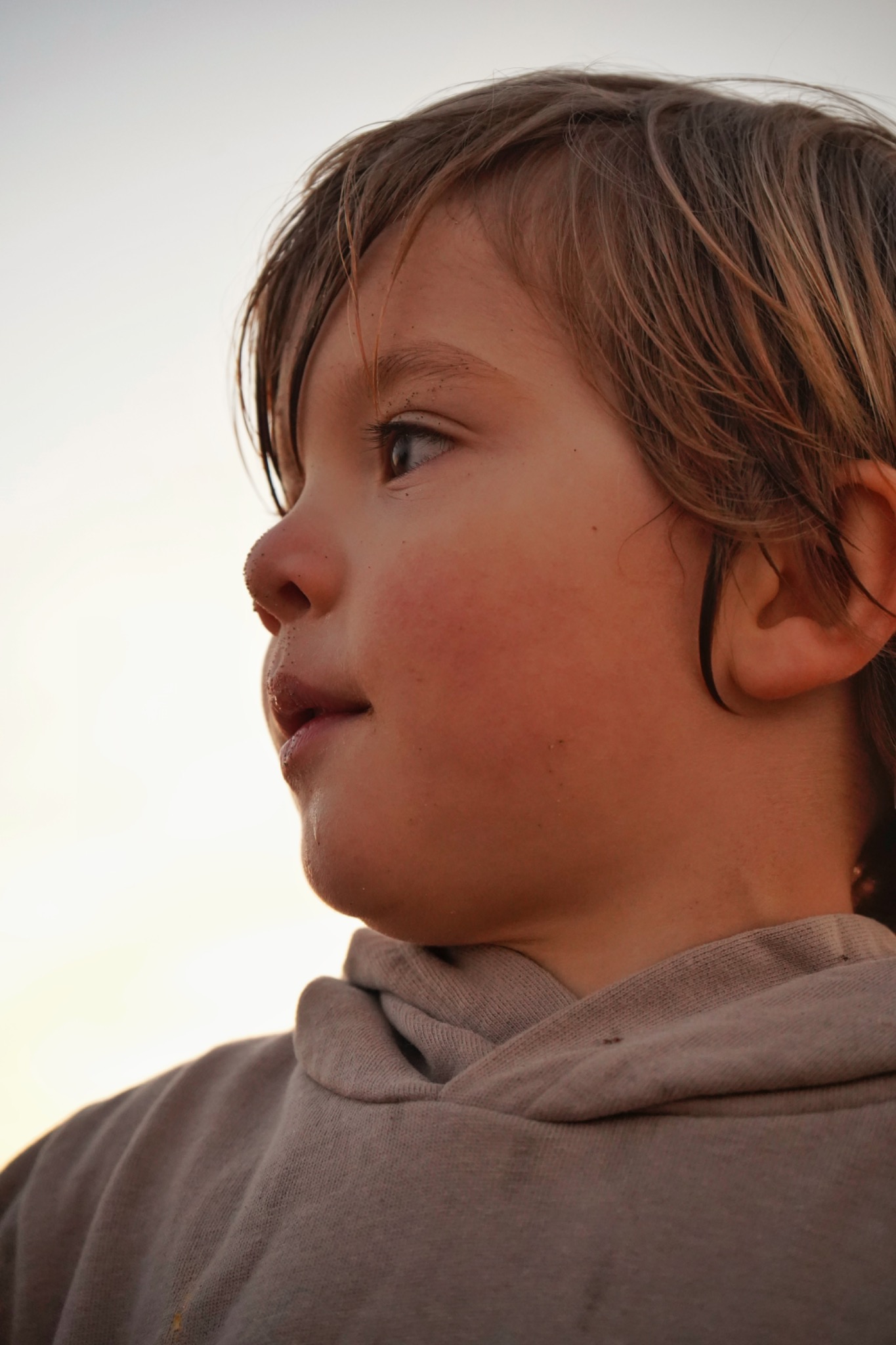 Profil młodego chłopca z piaskiem na nosie, ubrany w szarą bluzę z kapturem, spoglądający w dal, oświetlony ciepłym światłem.