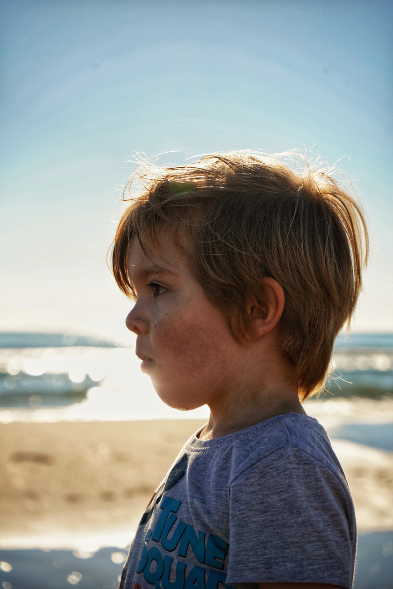 Profil chłopca z piegami na plaży, z kroplą wody na policzku, w tle morze i jasne niebo.