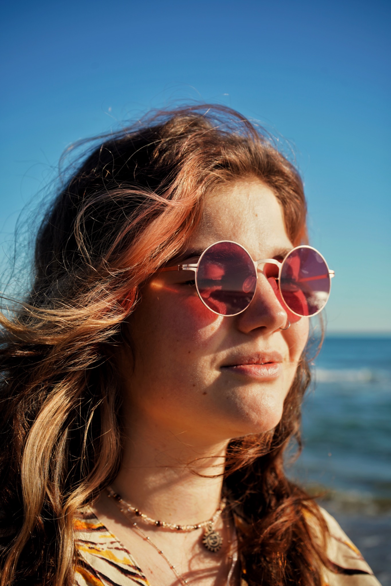 Portret kobiety w okularach przeciwsłonecznych z różowymi szkłami, stojącej na tle błękitnego nieba i morza, z włosami z pasemkami w odcieniach różu i blondu, oraz delikatną biżuterią na szyi.