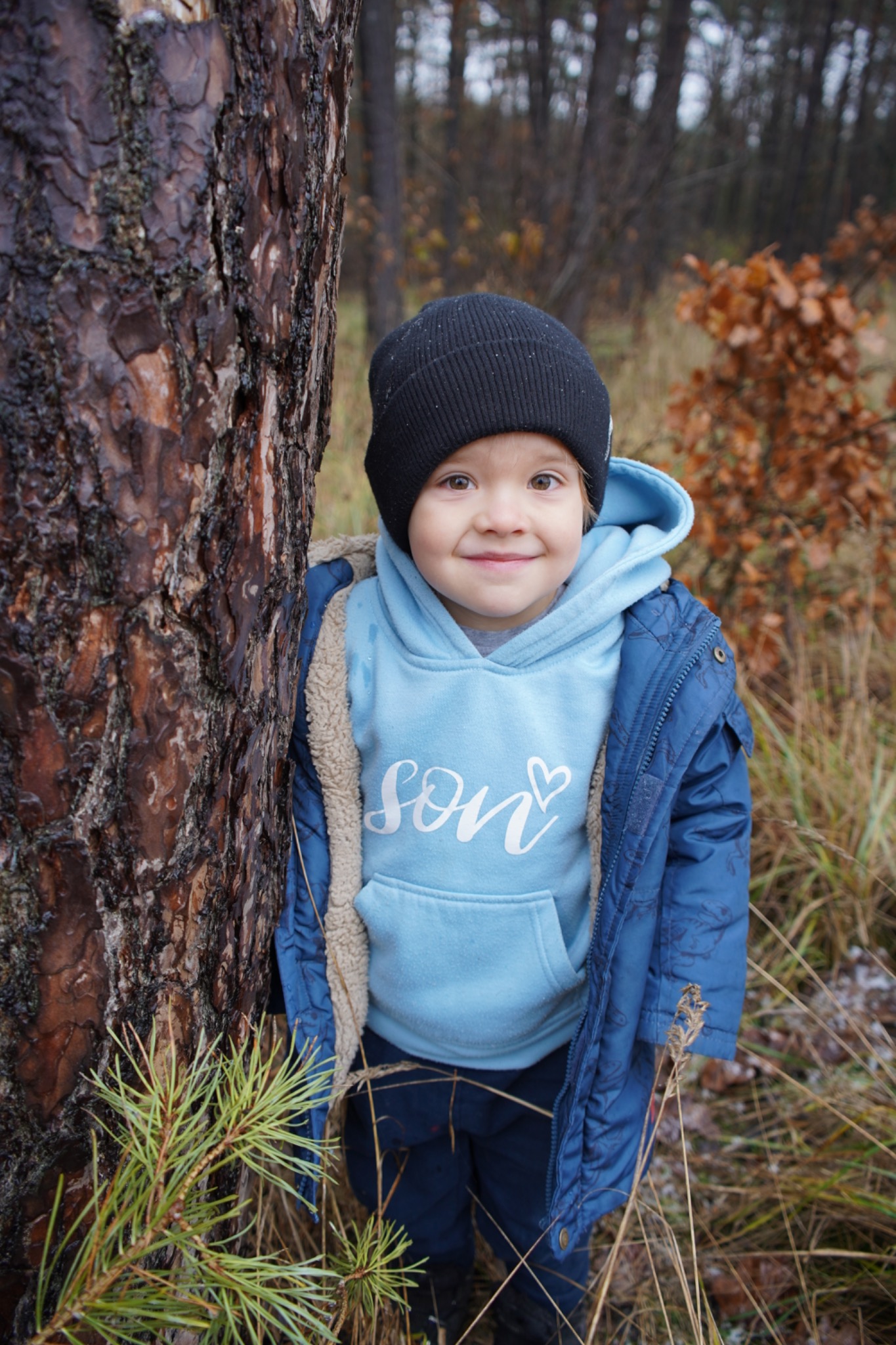 Uśmiechnięty chłopiec w czapce i niebieskiej bluzie z kapturem, oparty o sosnę w Pniewach, w tle jesienny las.