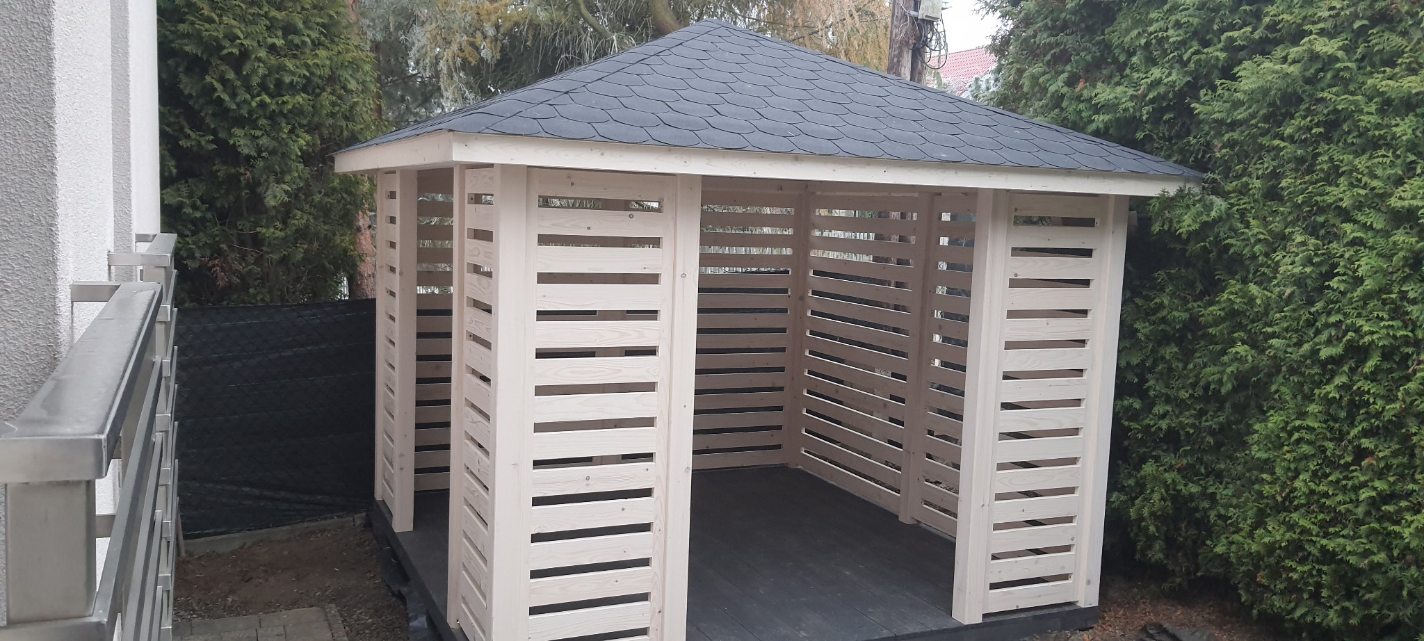 Nowoczesna, jasna altana z drewna z poziomymi szczelinami w ścianach i ciemnym, łupkowym dachem, otoczona zielenią.