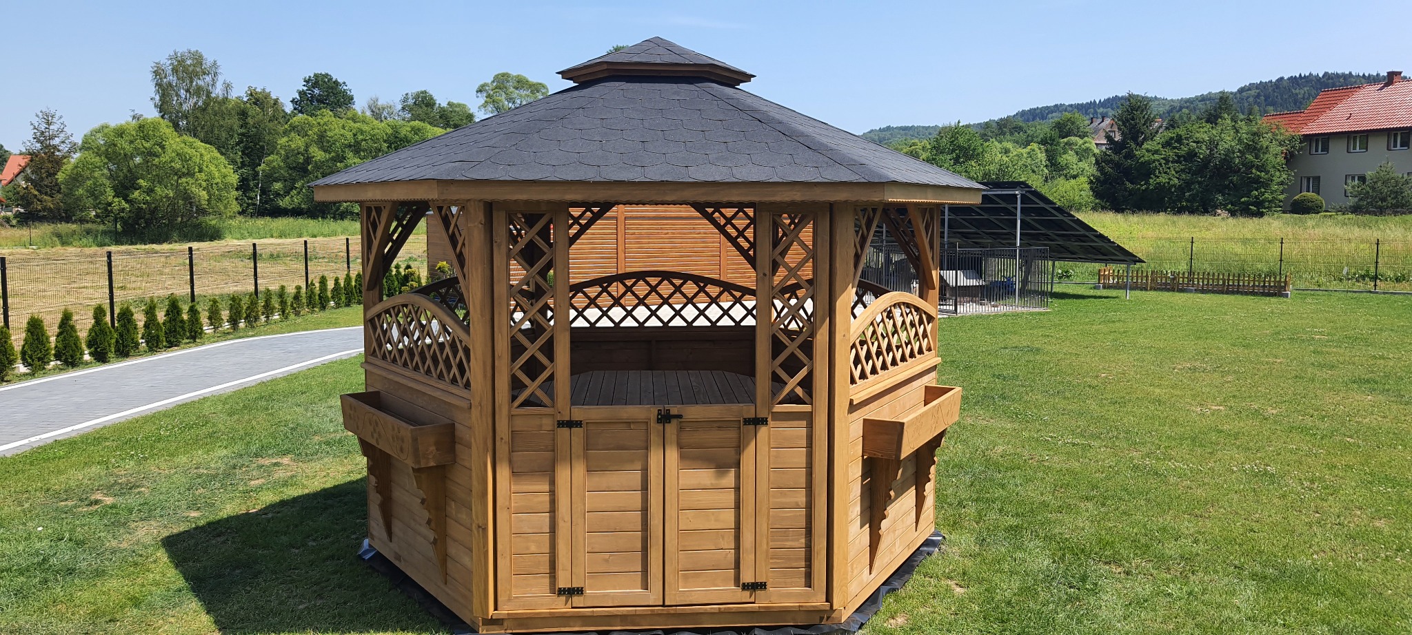 Ośmioboczna, drewniana konstrukcja z ciemnym, spadzistym dachem pokrytym gontem bitumicznym, ozdobnymi kratownicami i donicami, ustawiona na zielonym trawniku w ogrodzie, z widokiem na drzewa...