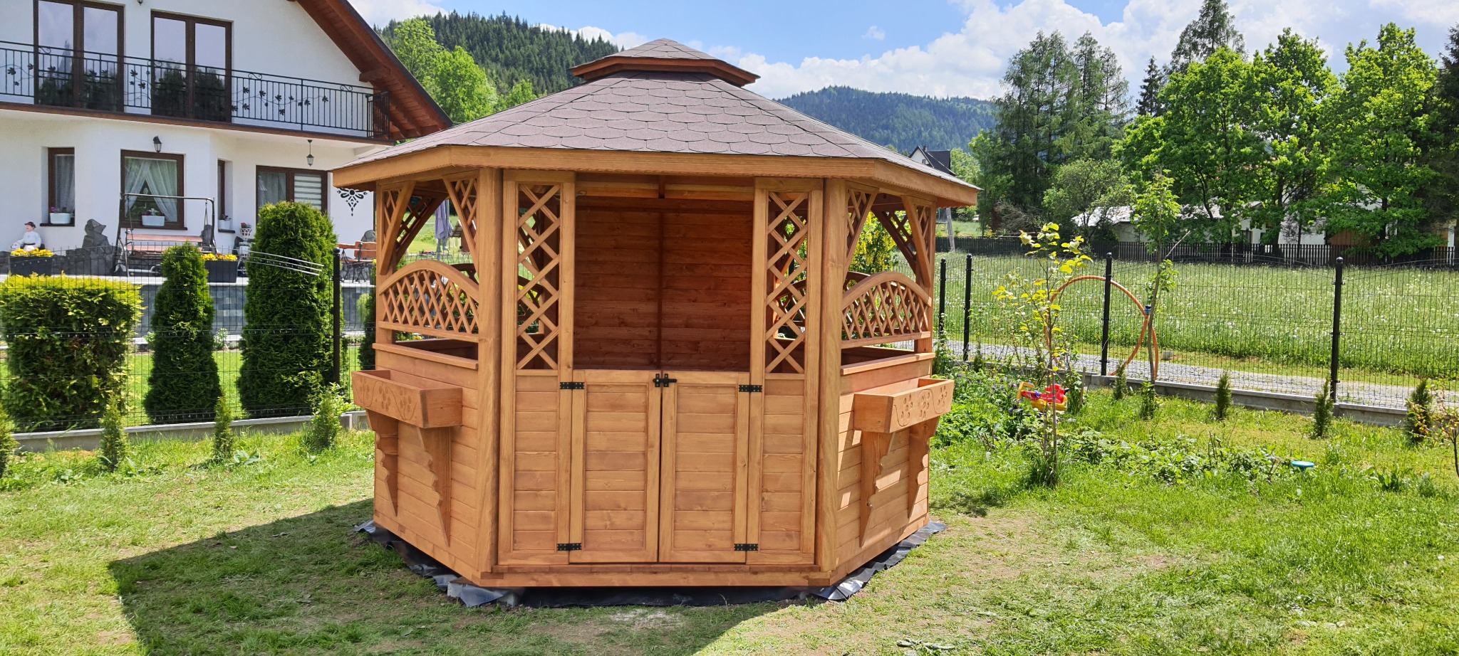 Ośmiokątna, drewniana altana z brązowym dachem, ozdobnymi kratownicami i donicami, ustawiona na trawniku przed domem w słoneczny dzień.