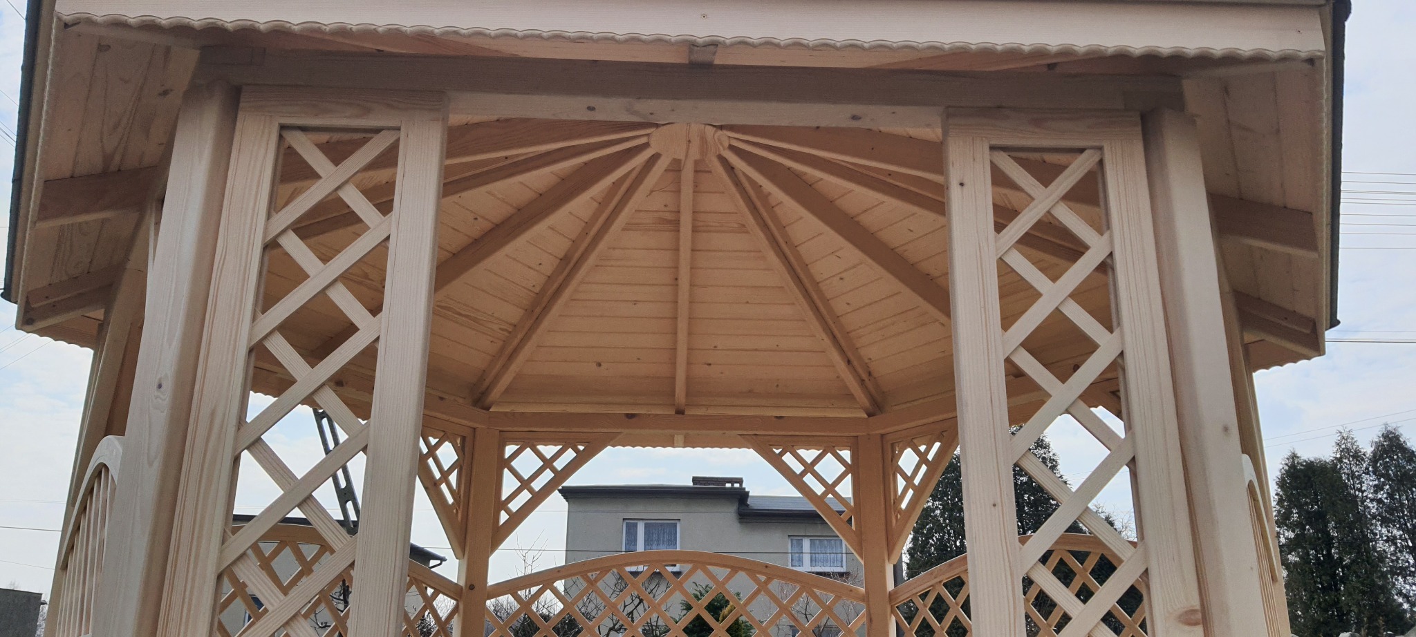 Wnętrze drewnianej altany z ażurowymi ścianami w Makowie Podhalańskim, widok na dach i fragment otoczenia.