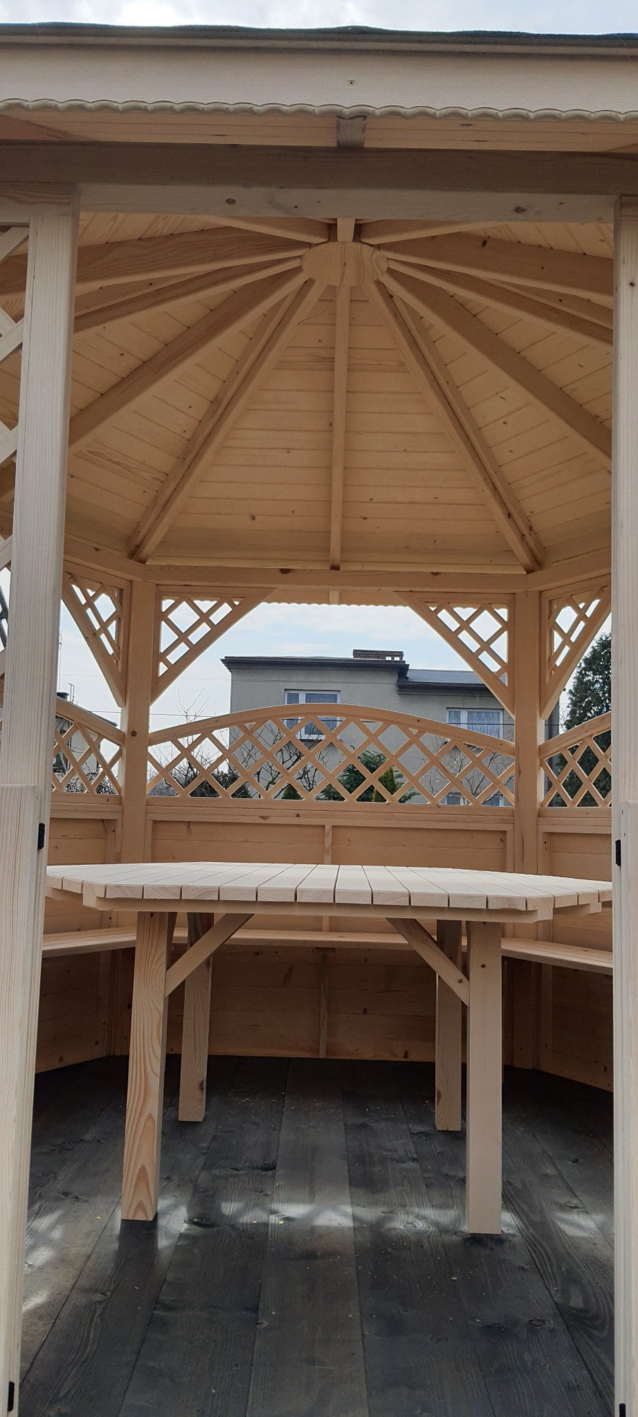 Wnętrze nowej, jasnej, drewnianej altany z ławkami i stołem, widok na budynek mieszkalny przez ażurowe ściany.