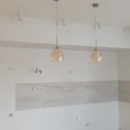 Wnętrze kuchni w stanie deweloperskim z białymi ścianami, jasnymi płytkami nad blatem, widocznymi gniazdkami elektrycznymi, otworami na instalacje i dwoma lampami wiszącymi z beżowymi kloszami.