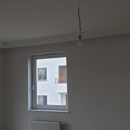 Dekor bud - Świeżo pomalowane szare ściany w pokoju z widokiem na zewnątrz, widoczny grzejnik pod oknem, wisząca lampa i żarówka na kablu.
