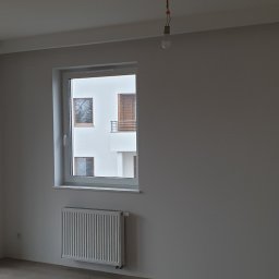 Dekor bud - Wnętrze pokoju po malowaniu na szaro, widoczny grzejnik, okno z widokiem na budynek i balkon z deskami oraz drzwi balkonowe. Na suficie wisi żarówka bez klosza.