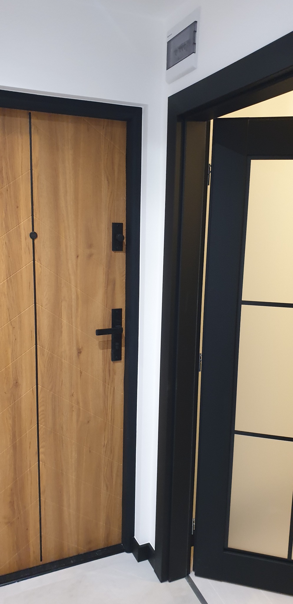Dwa rodzaje drzwi wewnętrznych: drewniane z czarnymi detalami i szklane z czarną ramą, na tle białej ściany z widoczną skrzynką elektryczną.