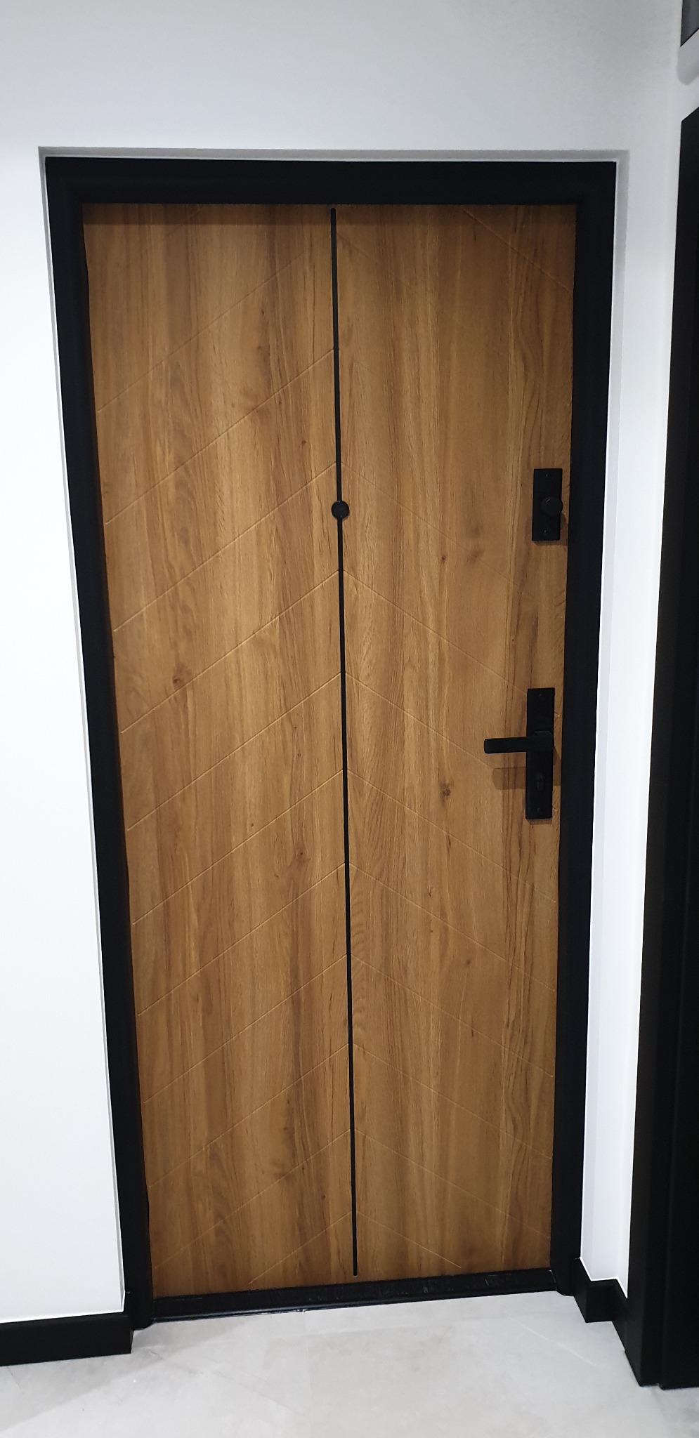 Drewniane drzwi wejściowe z czarną ramą i pionowym podziałem, wzór chevron na panelach, zamontowane w białej ścianie.