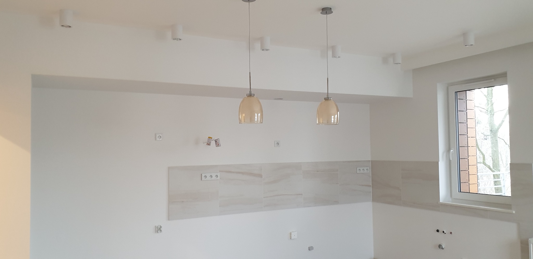 Wnętrze kuchni w stanie deweloperskim z białymi ścianami, jasnymi płytkami nad blatem, widocznymi gniazdkami elektrycznymi, otworami na instalacje i dwoma lampami wiszącymi z beżowymi kloszami.