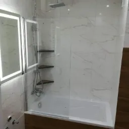 Nowoczesna łazienka z wanną obudowaną drewnopodobnymi panelami, przeszklonym parawanem nawannowym i marmurowymi płytkami ściennymi. Dodatkowo widoczne podświetlane lustro i narożne półki w kabinie...