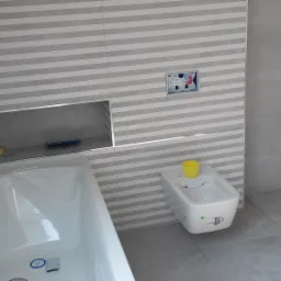 Wnętrze łazienki w trakcie wykańczania: biała wanna, bidet z żółtym kubkiem, szare płytki w paski na ścianach i podłodze, widoczny odpływ liniowy oraz wnęka w ścianie.