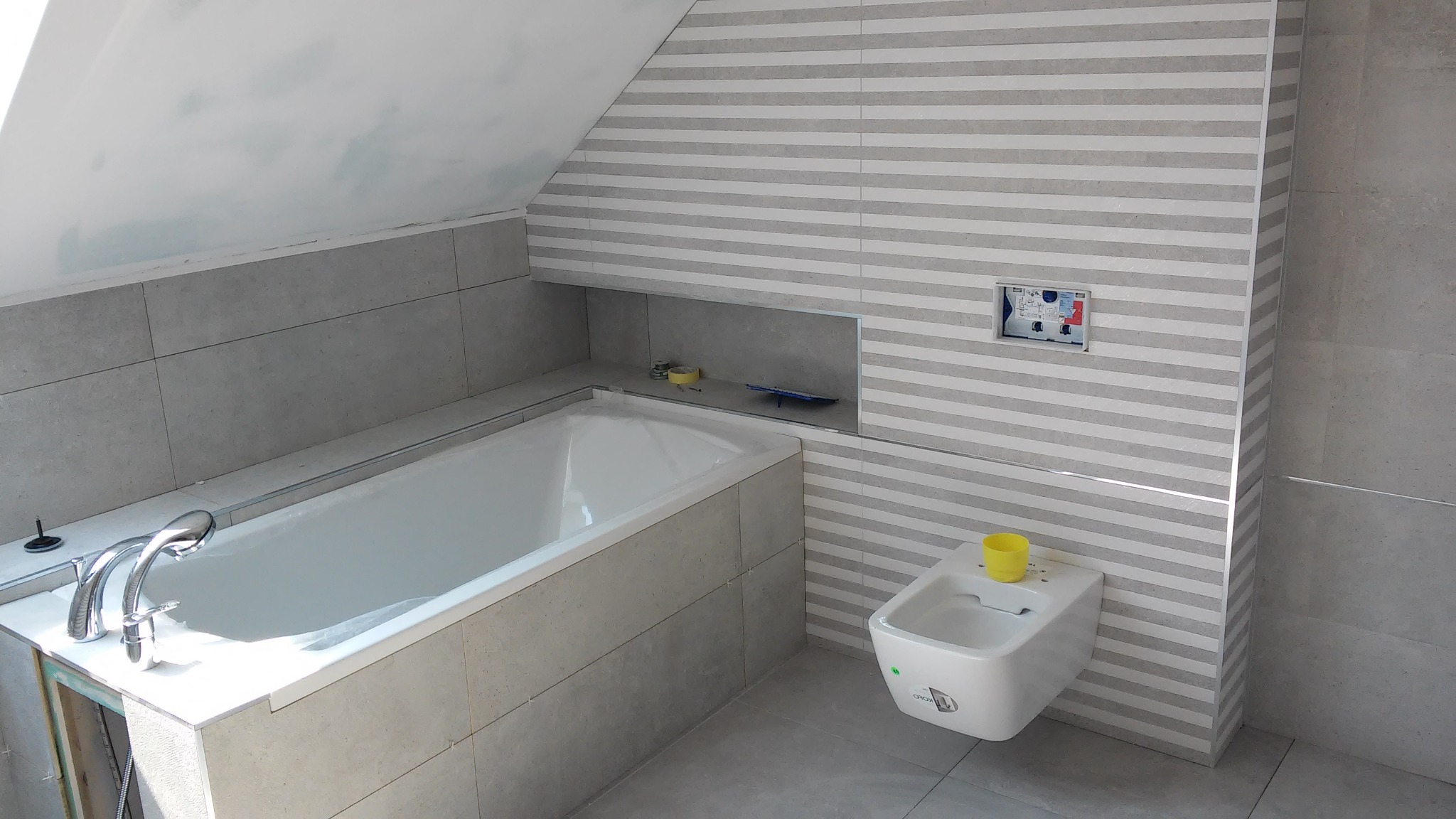 Łazienka na poddaszu z wanną obudowaną szarą płytką, białą armaturą i ścianami wyłożonymi płytkami w poziome pasy. W ścianie wnęka z akcesoriami i zamaskowany panel sterujący spłuczką.