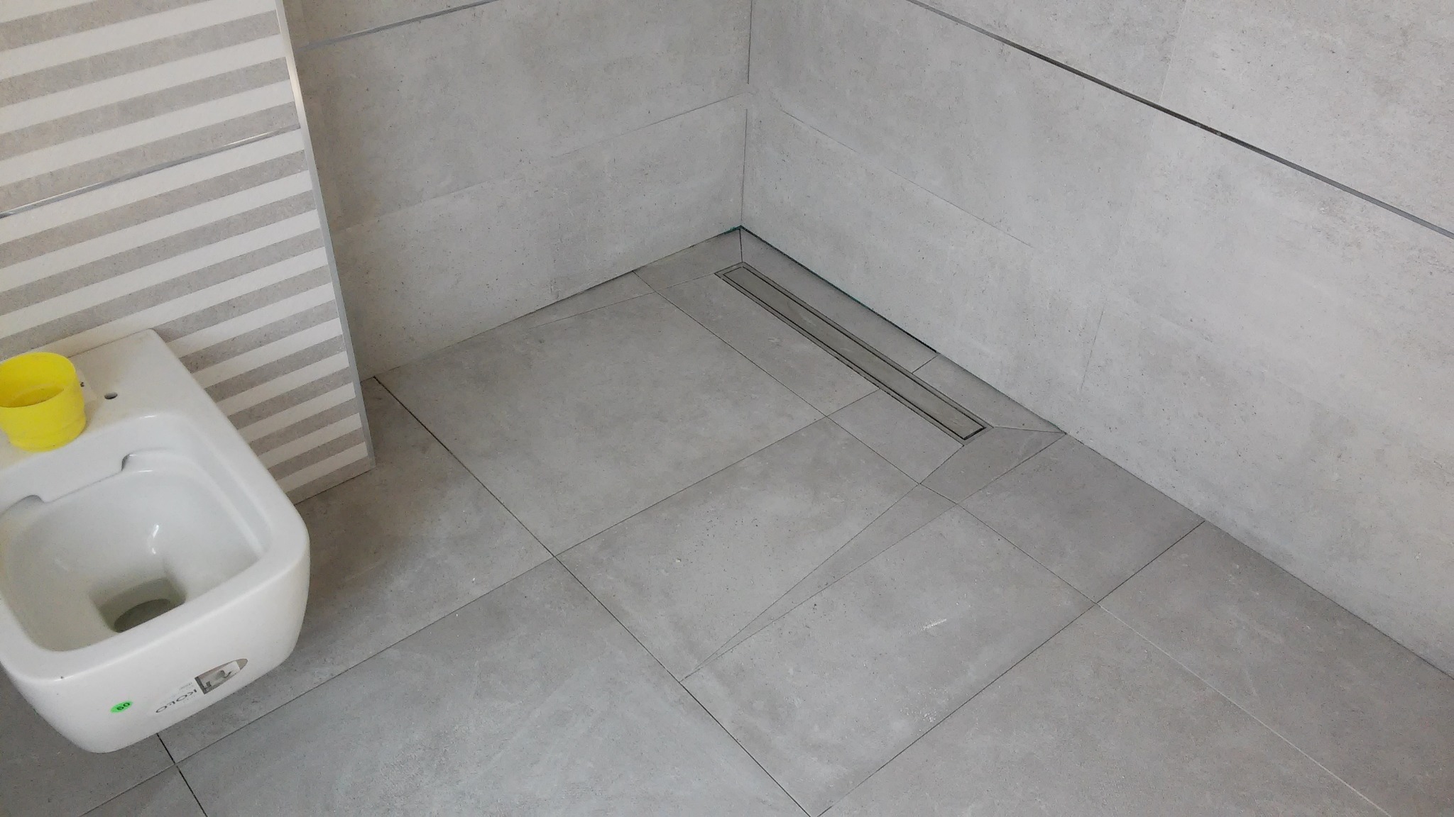 Szare płytki podłogowe i ścienne w łazience z odpływem liniowym i wiszącą toaletą z żółtym kubkiem.