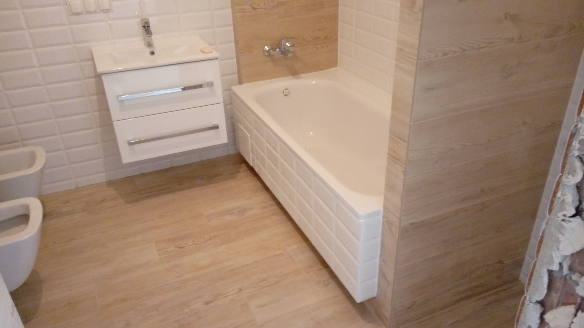 Nowoczesna łazienka z wanną obudowaną białymi kafelkami, umywalką z szafką w kolorze białym, bidetem i podłogą imitującą drewno, częściowo wykończona, z widocznymi fragmentami surowej ściany.