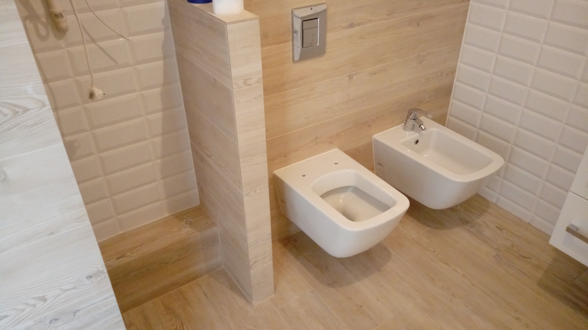 Nowoczesna łazienka z podwieszaną toaletą i bidetem, wykończona płytkami imitującymi drewno i białymi płytkami w kształcie cegiełek.