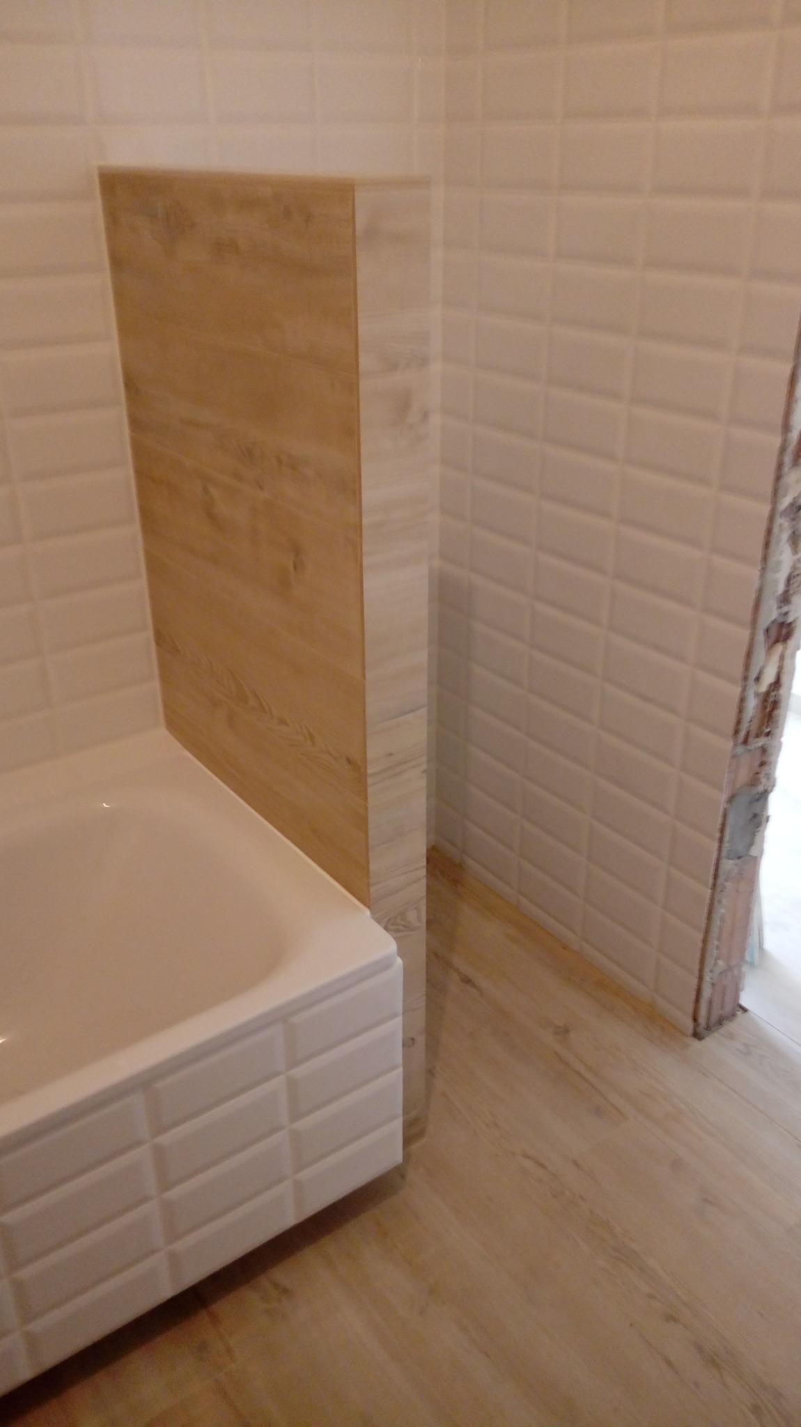 Wykończona łazienka z wanną obudowaną kafelkami w kształcie cegiełek, ściany wyłożone białymi płytkami, a podłoga panelami imitującymi drewno.