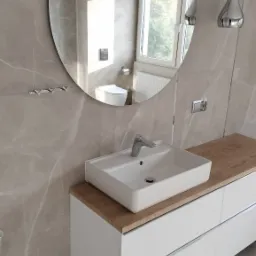 Nowoczesna łazienka z umywalką nablatową na białej szafce z drewnianym blatem, okrągłe lustro odbija toaletę i okno, ściany wyłożone płytkami imitującymi marmur.
