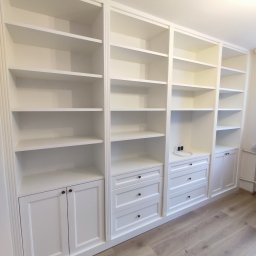 SZTANI Stanisław Mizdal - Biała zabudowa meblowa z szafkami i szufladami, tworząca biblioteczkę z pustymi półkami, z delikatnymi zdobieniami frezowanymi na obrzeżach.