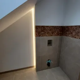 Nowoczesna łazienka na poddaszu z ukośną ścianą i oświetleniem LED wzdłuż krawędzi, brązowa mozaika nad szarymi płytkami, widoczny stelaż podtynkowy.