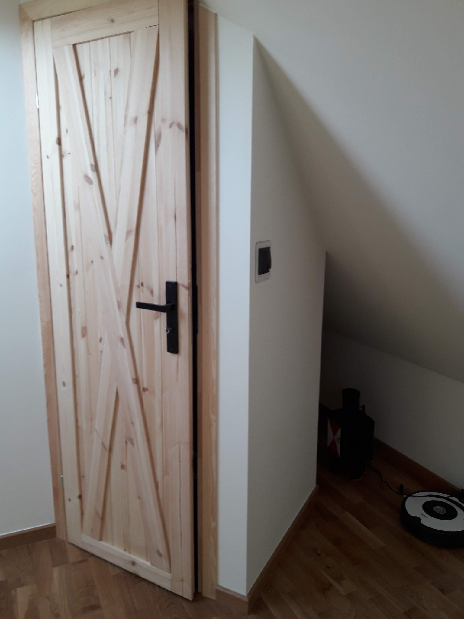 Drewniane drzwi w stylu rustykalnym z czarną klamką, zamontowane w białej ścianie na poddaszu, w tle widoczny odkurzacz automatyczny i pojemnik.