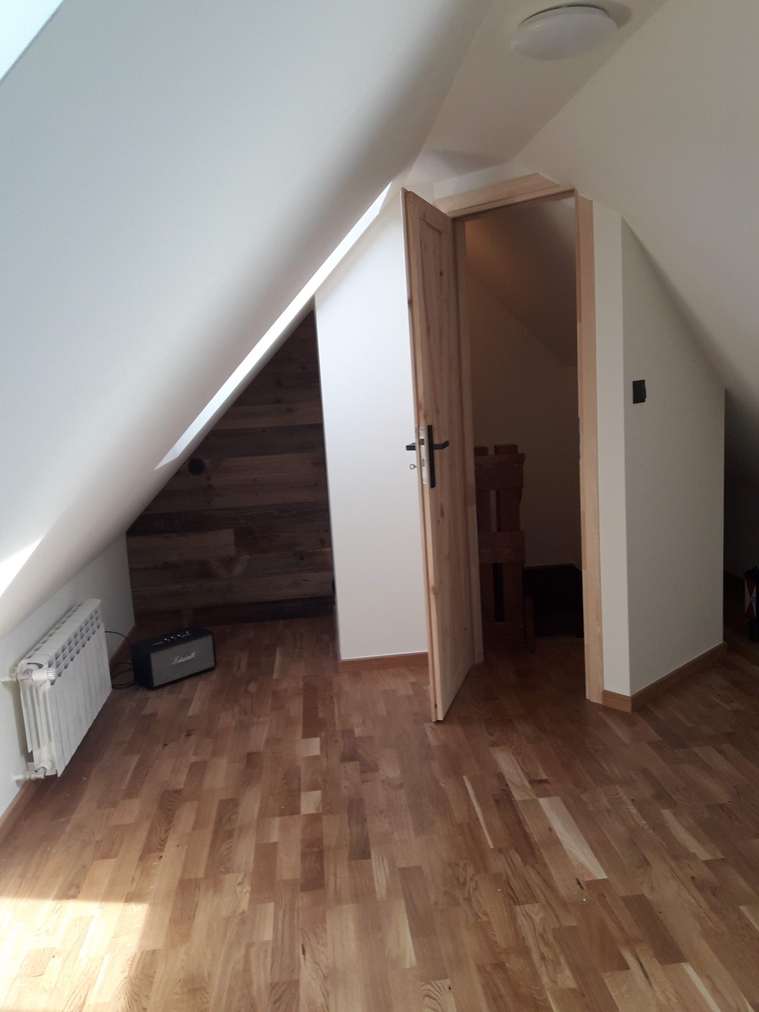 Poddasze po remoncie: skosny sufit, drewniana podłoga, grzejnik, wzmacniacz Marshall i otwarte drzwi do garderoby z drewnianą ścianą.