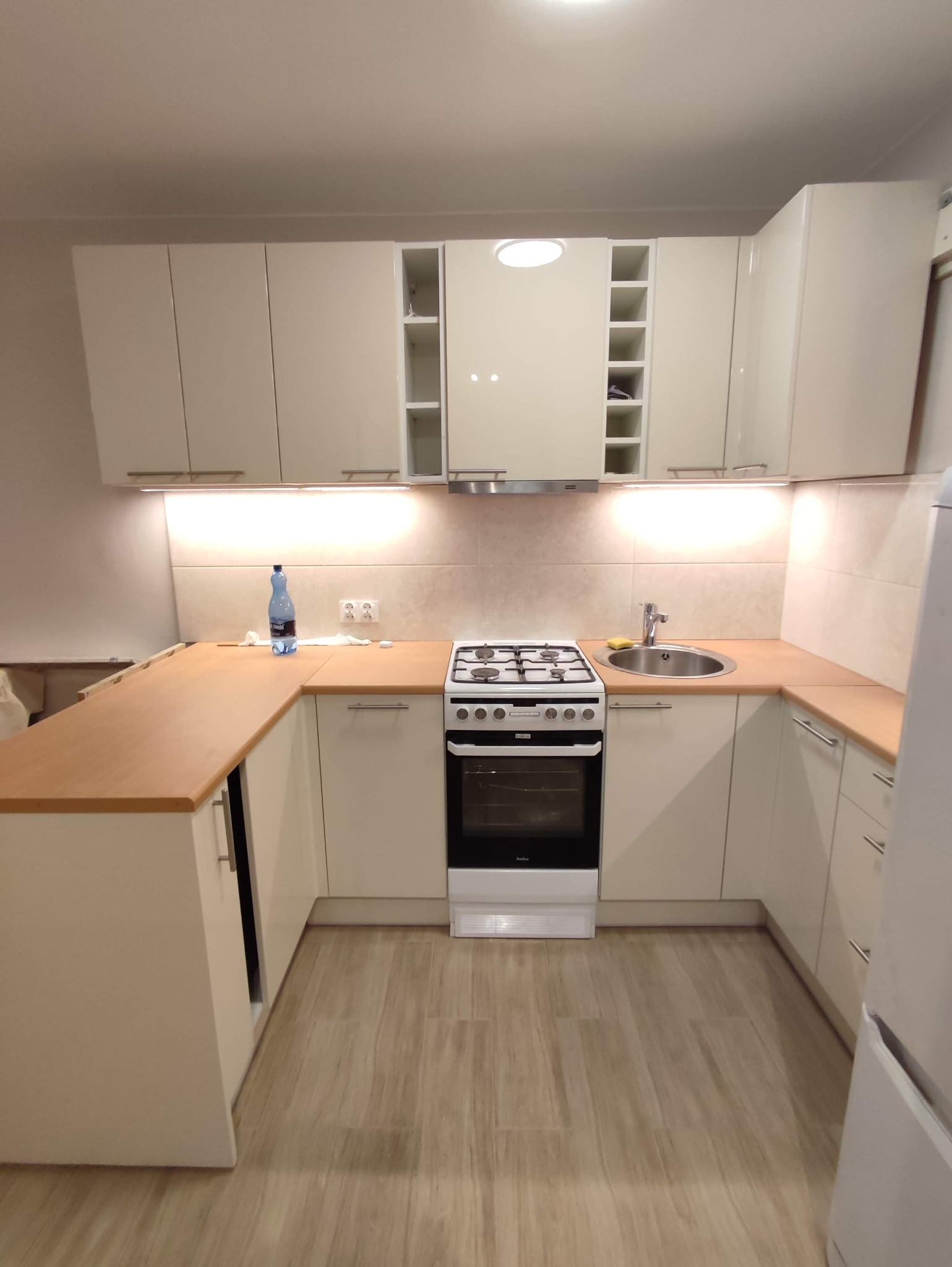 Jasna kuchnia z drewnianym blatem, białymi szafkami i sprzętami AGD, oświetlona listwą LED pod szafkami wiszącymi.