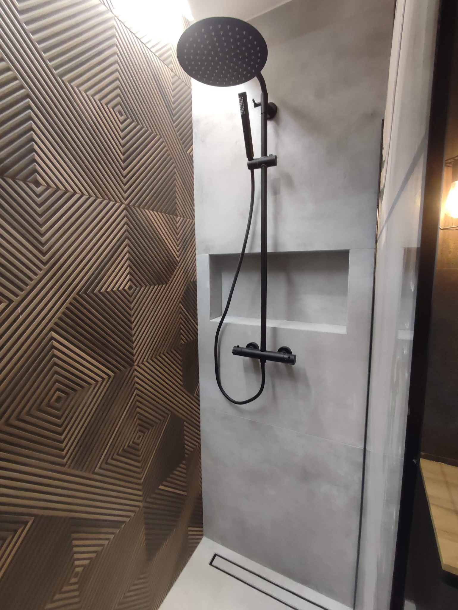 Nowoczesna kabina prysznicowa z deszczownicą w kolorze czarnym, ściana z dekoracyjnymi panelami 3D o wzorze geometrycznym w odcieniach brązu i beżu, oraz szara ściana z wnęką.